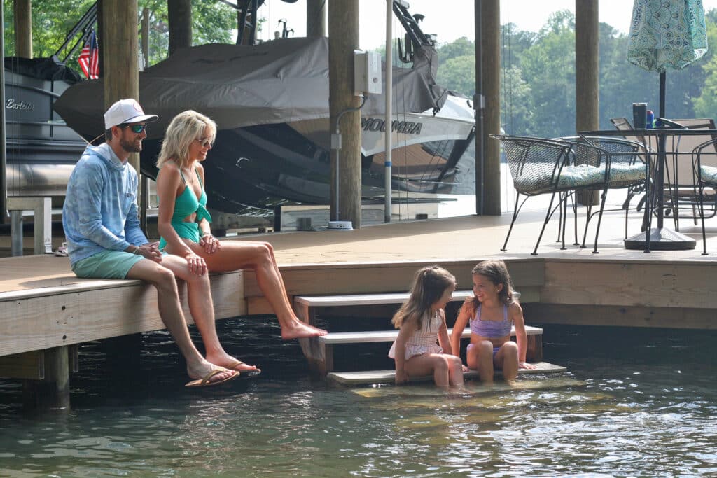 Parents relaxing on the edge of their stationary dock with their two daughters sitting on the water steps, enjoying the beautiful lakeside view.