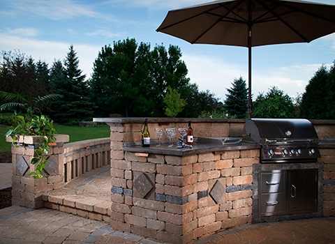Beautiful outdoor kitchen with a bar, built-in grill, ambient lighting, sun umbrella, and a stunning landscaped backdrop.