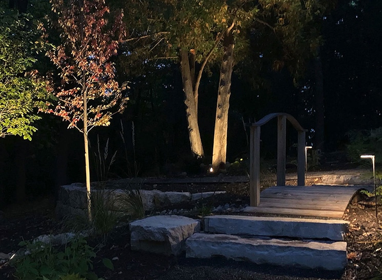 Beautifully lit garden pathway with trees and stone bridge illuminated by landscape lighting at night.
