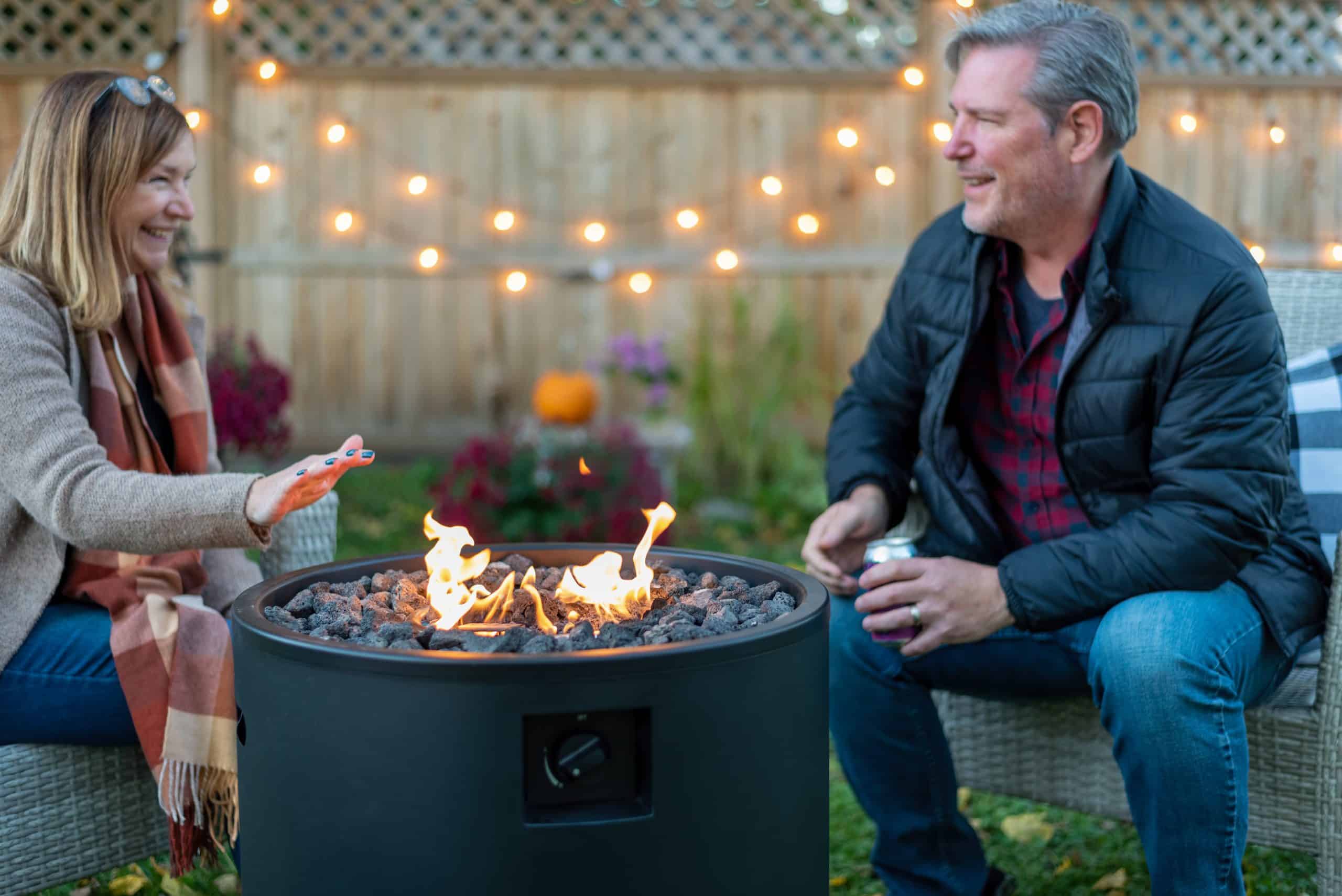 Middle-aged couple sitting by a compact fire pit in a small backyard, enjoying a cozy evening with string lights in the background.